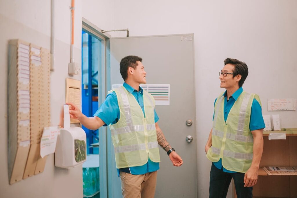 Image showing two nonexempt employees in safety vests clocking in at a workplace time tracking station.